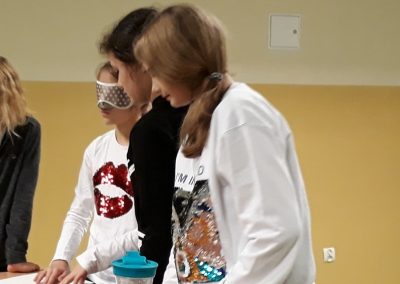 trzy uczennice przy ławce. Jedna ma zasłonięte oczy i poznaje braila, druga patrzy na nią, a trzecia patrzy na kostke rubika. przed nimi są szklanki z wodą oraz karafka
