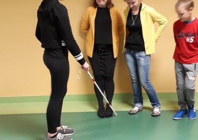 na zdjęciu 4 osoby: nauczycielka i troje uczniów, jedna uczennica ma w ręce laskę dla niewidomych, nauczycielka z resztą uczniów stoi pod ścianą